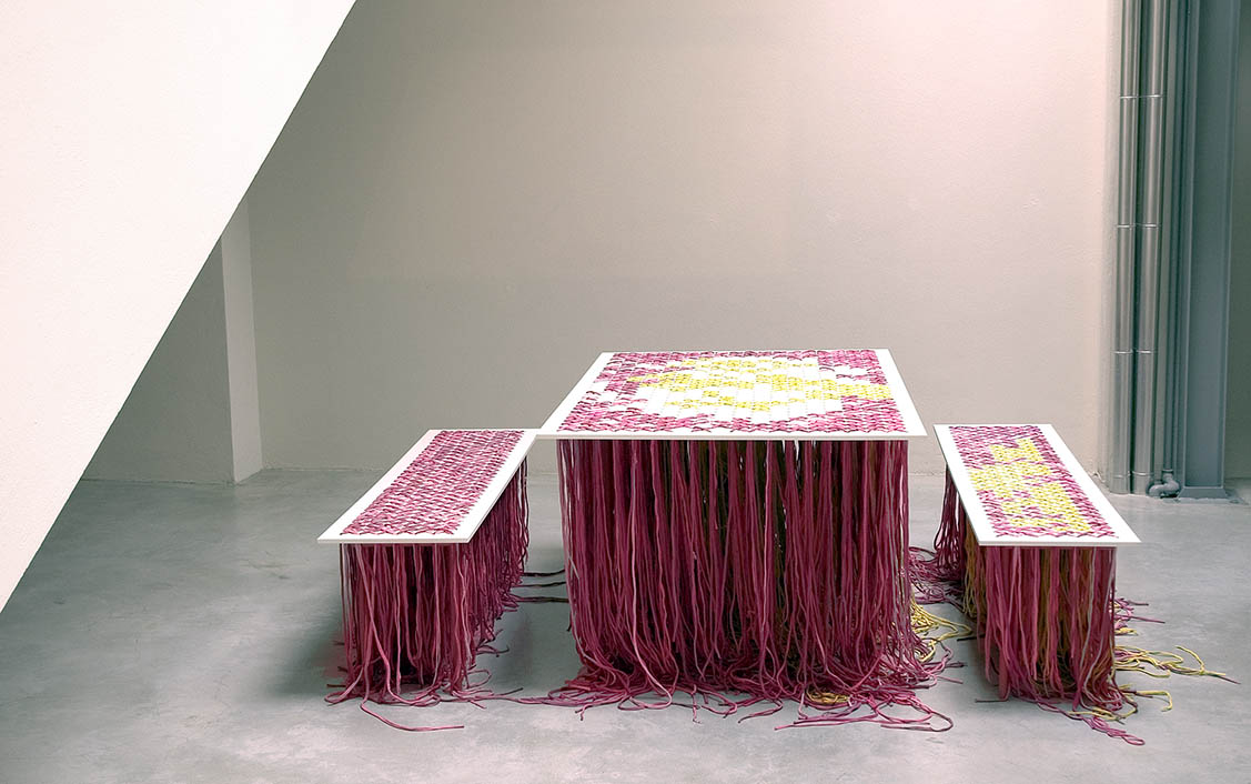 A table with hundreds of coloured threads hanging down from it so that you can't see what's underneath it.