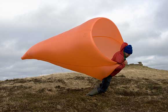 A man holds a large orange fabric funnel like a wind sock, it is filled by the wind and he's fighting to stand upright.